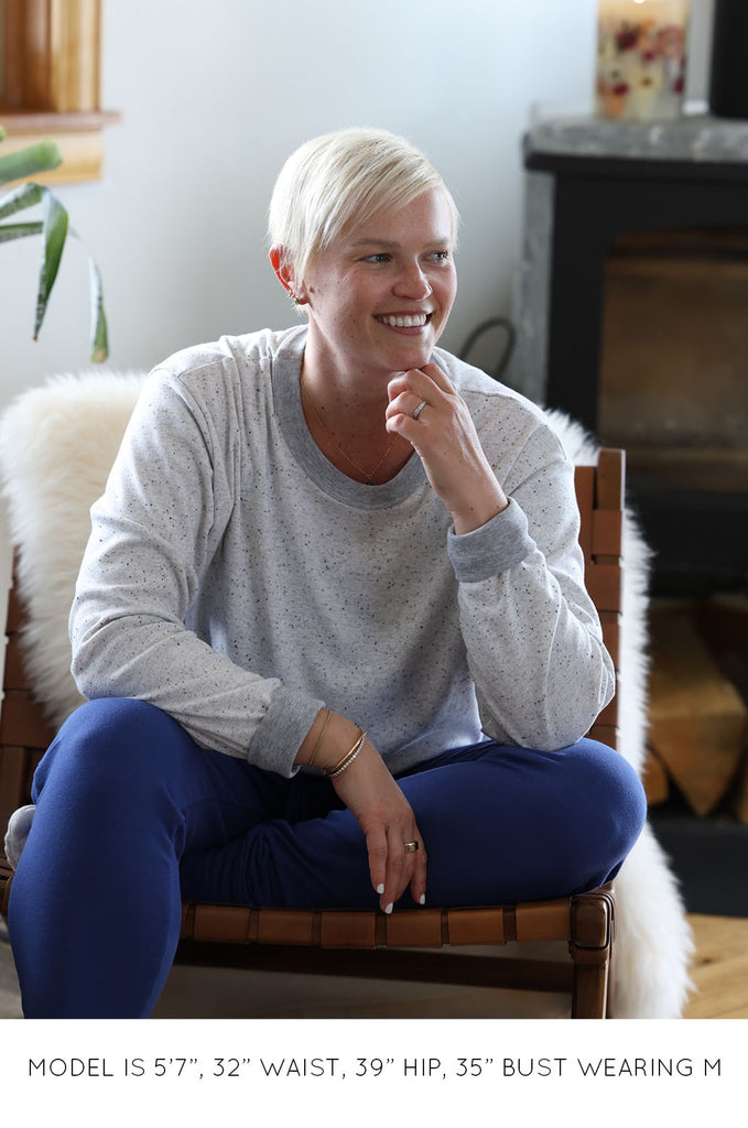 Model sitting in chair wearing blue track pants and grey long sleeve Miakoda top. 