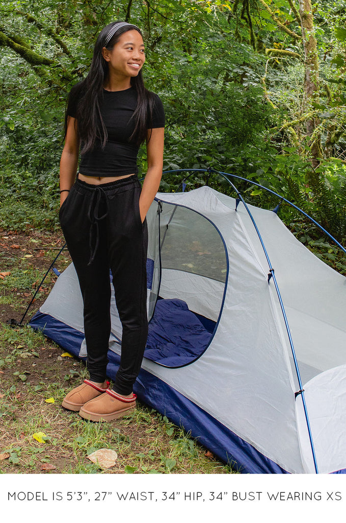 Model standing in front of tent wearing a black Miakoda Baby Tee with black pants. 