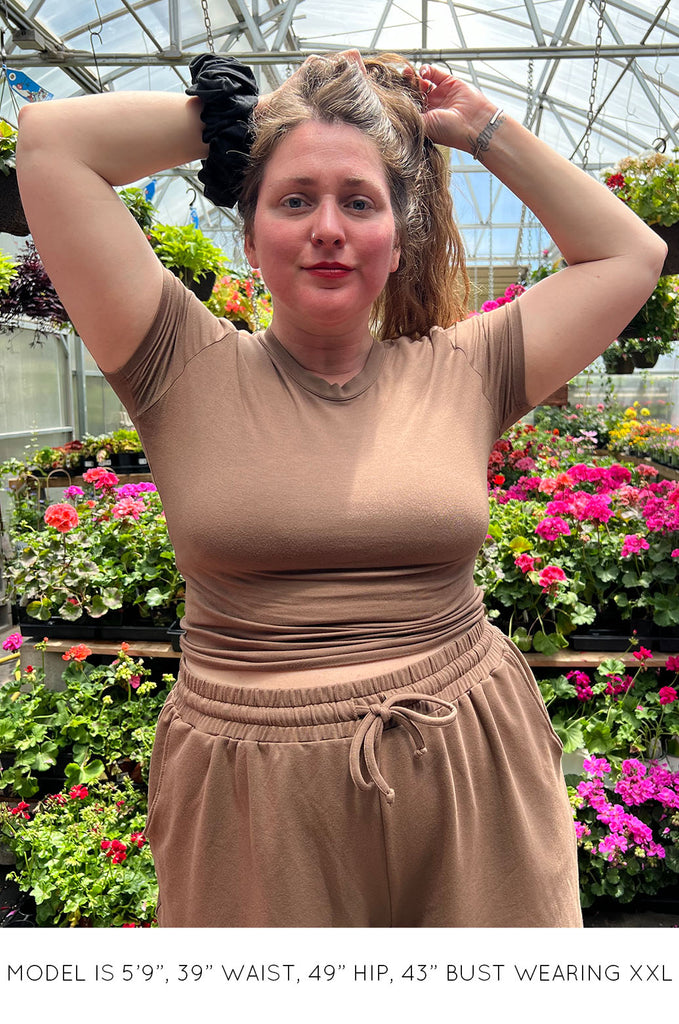Model standing in greenhouse wearing brown Miakoda Baby Tee with matching brown shorts. 