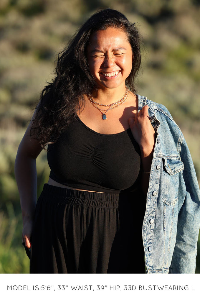 Model smiling in the sun, wearing black Miakoda Brami crop top with black pants and denim jacket. 