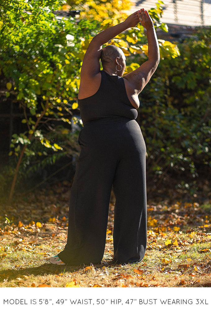 Back view of model in front of tree wearing black racer back tank and black wide leg pants by Miakoda. 